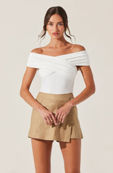 A woman is wearing an off-the-shoulder white top with draped and layered fabric details across the bust and shoulders. She is also wearing high-waisted beige shorts with a slight A-line shape, a front button closure, and side pockets.